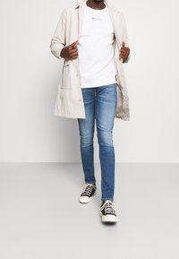 Giacca beige leggera sopra una felpa bianca, abbinata a jeans blu aderenti e sneaker nere con suola bianca.