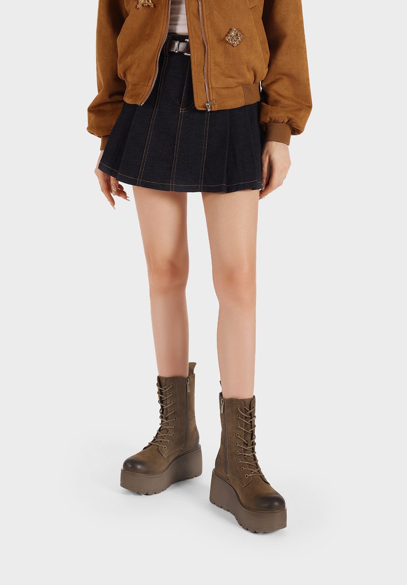 Stivali platform marroni con lacci, finitura in pelle texturizzata e suola spessa, abbinati a una gonna di jeans e a una giacca bomber marrone.