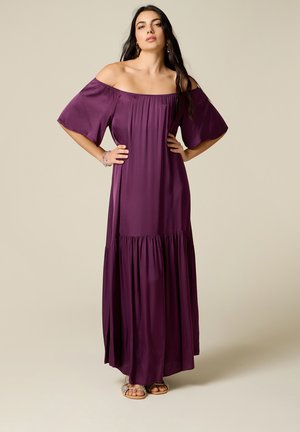Frau trägt ein violettes schulterfreies Maxikleid mit weiten Ärmeln, steht mit den Händen in der Hüfte vor einem schlichten Hintergrund.