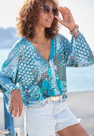 Blue and turquoise patterned blouse with long sleeves, deep V-neck, and lightweight fabric, paired with light denim shorts and a gold belt.