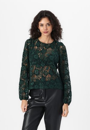 Haut en dentelle verte à manches longues avec des motifs floraux et un col rond, associé à un pantalon en cuir noir. La dentelle présente une texture transparente.