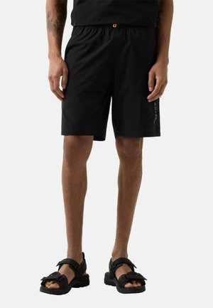 Mann trägt schwarze Sportshorts mit seitlichem Logo und schwarze Sandalen mit offenem Zehenbereich, steht mit entspannten Händen seitlich am Körper vor einem neutralen Hintergrund.