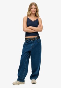Pantaloni a gamba larga in denim blu, cintura nera con borchie, top camicia navy con bordi in pizzo e scarpe da ginnastica chiare. La modella è in piedi con le braccia incrociate.