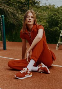 Conjunto deportivo rojo con cuello con cremallera, combinado con zapatillas rojas y blancas, y calcetines blancos hasta el tobillo, sentado en una cancha color beige.