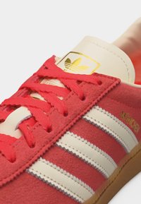Κόκκινο αθλητικό παπούτσι Adidas με λευκές ρίγες, κόκκινες κορδόνια, καουτσούκ σόλα και χρυσό λογότυπο Adidas στη μπεζ γλώσσα. Κείμενο "München" σε χρυσό στο πλάι.