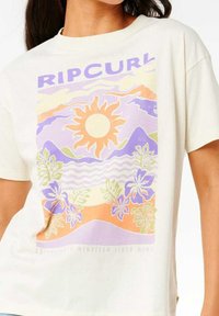 T-shirt couleur crème présentant un graphique coloré d'un soleil, de montagnes, de vagues et de fleurs tropicales dans des tons violets, oranges et verts.