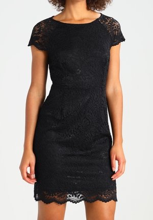 Mujer con un vestido corto ajustado de encaje negro con borde festoneado, de pie contra un fondo claro y liso.