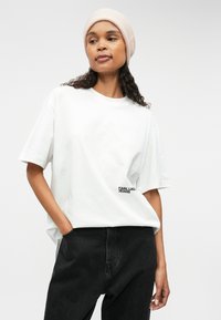 Λευκό βαμβακερό μπλουζάκι με στρογγυλή λαιμόκοψη και κοντά μανίκια, με το μαύρο λογότυπο "KARL LAG. JEANS" στην αριστερή πλευρά. Συνδυασμένο με μαύρο παντελόνι.