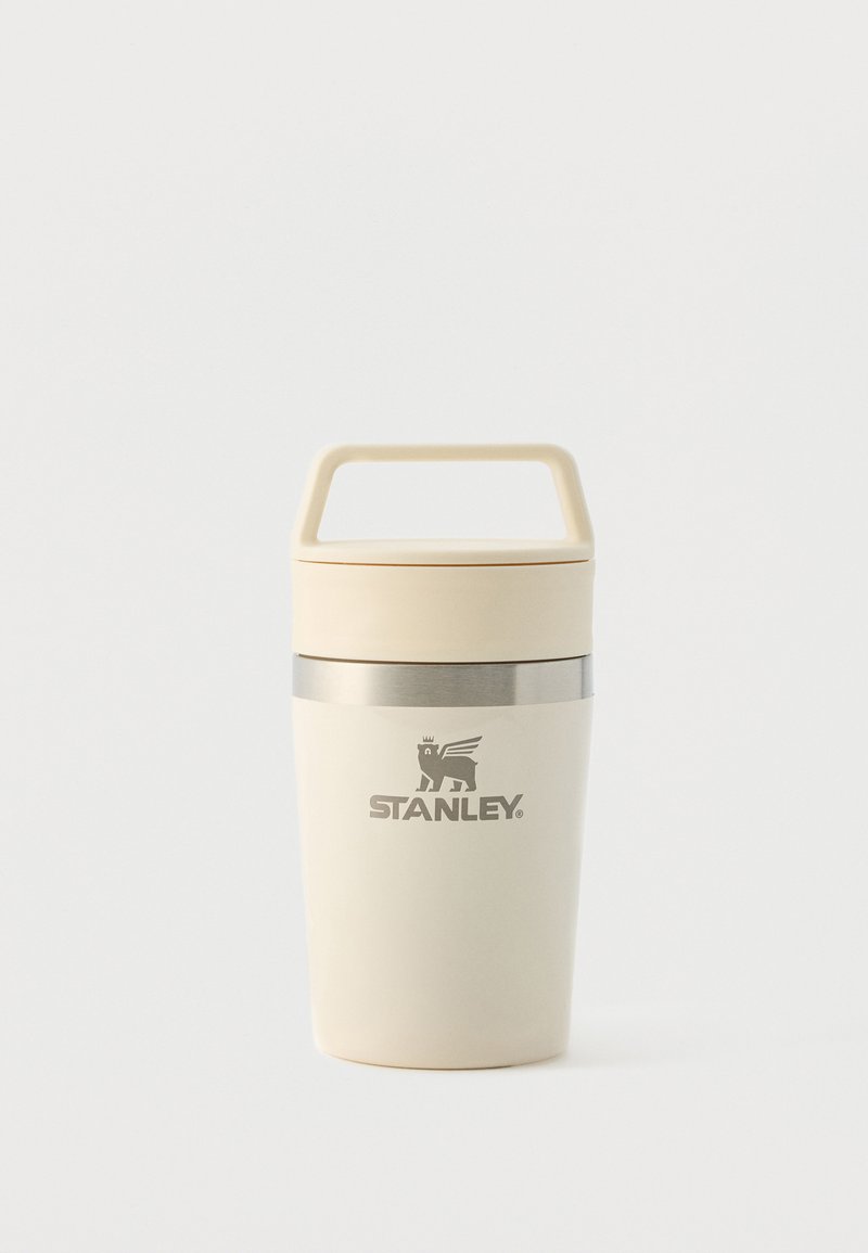 Cremefarbener Stanley Thermobecher mit Griff, Deckel und silbernem Ring nahe dem oberen Rand, vor einem schlichten weißen Hintergrund.