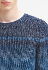 Sweter dzianinowy o teksturowanym wzorze w różnych odcieniach niebieskiego i ciemnego granatu. Okrągły dekolt i długie rękawy.