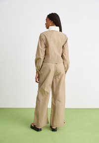 Jumpsuit beige, texturizado, ligero, de manga larga con puños elásticos, cuello alto y pantalones de pierna ancha, combinado con sandalias de plataforma negras.