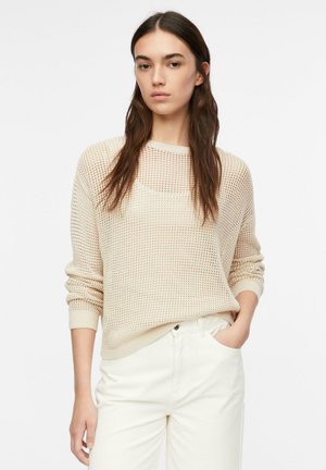 Junge Frau mit langen braunen Haaren, die einen beigen, offen gestrickten Pullover über einem Top und weißen, hoch taillierten Hosen trägt, steht mit einer Hand in der Tasche.