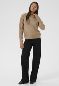 Lichtbruine, gestructureerde gebreide trui met ronde hals, gecombineerd met hoge zwarte wijde broek en zwarte schoenen. Effen achtergrond.