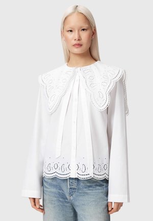 Kobieta z długimi blond włosami, ubrana w białą bluzkę z dużym koronkowym kołnierzem o falistym kształcie oraz dżinsy, stoi na jednolitym szarym tle.