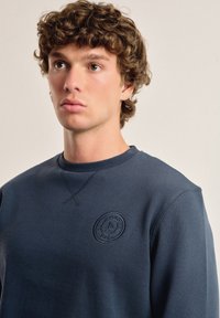 Sudadera azul marino con un logo bordado redondo en el pecho, cuello de canalé y tejido texturizado con un corte regular.