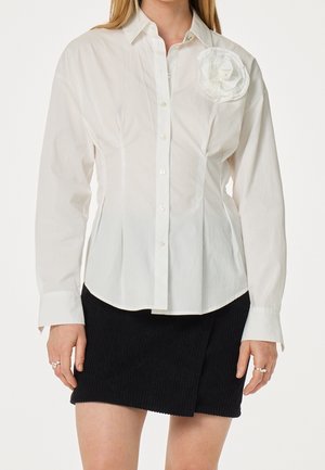 Femme portant une chemise blanche boutonnée avec une grande fleur en tissu sur le côté gauche de la poitrine et une mini-jupe noire texturée.