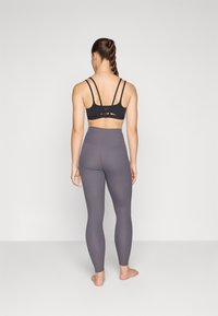 Soutien-gorge de sport noir avec plusieurs fines bretelles, associé à un legging taille haute en côte gris foncé, porté pieds nus.