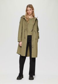 Oliwkowa parka z kapturem, z akcentami w postaci sznureczków, z kieszeniami naszytymi i z przodu na zamek, w połączeniu z zielonym topem i czarnymi spodniami. Czarne obuwie.