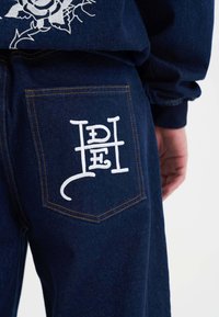 Ciemnoniebieskie dżinsy mają tylną kieszeń z białym haftowanym logo i kontrastującymi pomarańczowymi szwami. Materiał ma gładką teksturę.