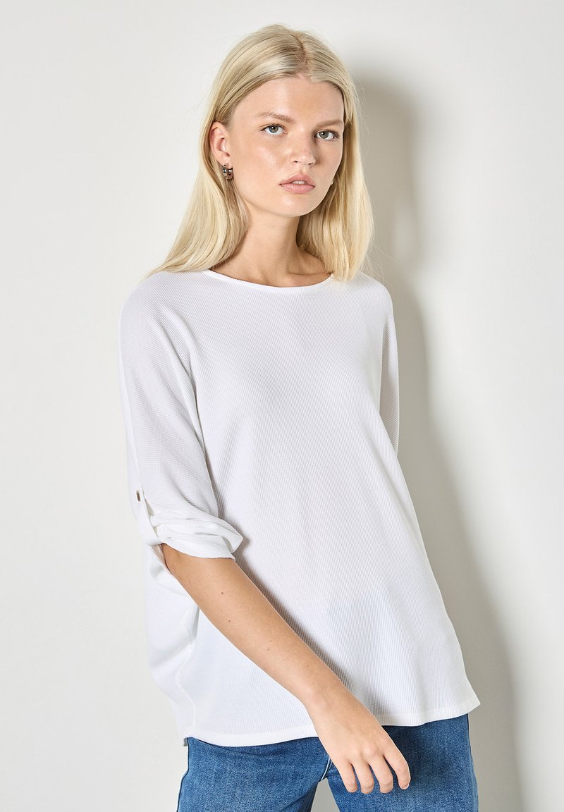 Pull blanc surdimensionné avec une surface texturée, encolure bateau et manches retroussées, associé à un jean bleu.