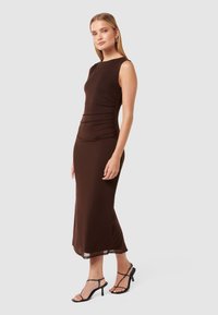 Robe marron sans manches avec un corsage ajusté et des détails froncés sur le côté, descendant jusqu'à mi-mollet. Portée avec des escarpins noirs à lanières.