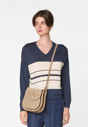 Vrouw die een blauw-wit gestreepte V-hals trui draagt, blauwe broek en een beige schoudertas, kijkt opzij tegen een witte achtergrond.