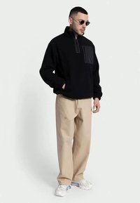 Homemem vestindo um fleece preto de meia-zíper, calças largas bege, tênis brancos e óculos de sol redondos, posando contra um fundo claro simples.