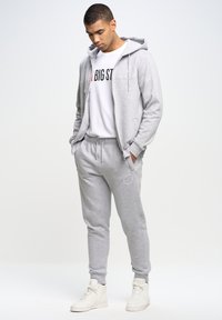 Grå hoodie med forlommer over en hvid t-shirt med sort tekst. Grå joggingbukser med elastiske manchetter. Hvide sneakers fuldender looket.