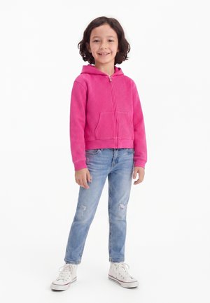 Barn står og smiler, iført en lys pink hættetrøje med lynlås, lyseblå revnede jeans og hvide høje sneakers.