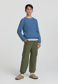 Niebieski sweter z dzianiny z ribbingu, nałożony na białą koszulę. Oliwkowe spodnie cargo z bocznymi kieszeniami oraz beżowe klapki o miękkiej teksturze.