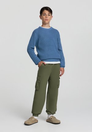 Maglione blu lavorato a maglia con costine, indossato sopra una camicia bianca. Pantaloni cargo verde oliva con tasche laterali e zoccoli beige dal tessuto morbido.