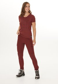 Bordeauxfarbene Sportbekleidung, bestehend aus einem figurbetonten T-Shirt mit dem Aufdruck "ADVENTURE" und einer schmal geschnittenen Hose mit Reißverschlusstaschen und Kniepaneelen.