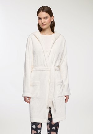 Donna che indossa un accappatoio bianco con cappuccio e cintura in vita, una maglia bianca sotto e pantaloni neri con motivi a cuore rosa e bianchi.