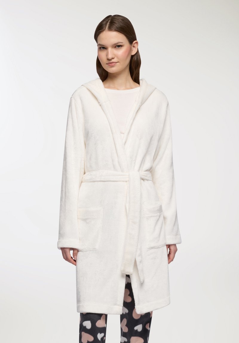 Donna che indossa un accappatoio bianco con cappuccio e cintura in vita, una maglia bianca sotto e pantaloni neri con motivi a cuore rosa e bianchi.