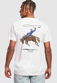 Hvit bomull T-skjorte med et grafisk motiv av en cowboy som rir på en brun hest, med teksten "LEVE I ØYEBLIKKET" i svart under bildet.