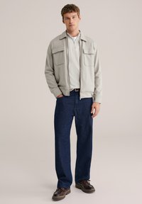 helehall zip-up jakk kahe esimest taskutega, kantud valge särke kohal, kombineeritud tumeliste siniste teksade ja pruunide ketskingadega.