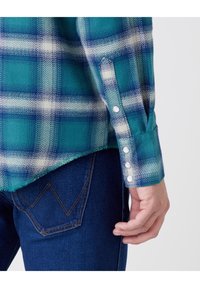 Heren flanellen overhemd in turkoois en blauwe ruitpatroon, met witte accenten, knoopmanchetten en een iets gebogen zoom. Donkerblauwe jeans zichtbaar.