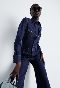 Salopetă din denim cu spălare închisă, dotată cu două buzunare la piept, cusături albe și manșete cu butoane. Modelul ține o geantă de umăr de culoare albastră deschisă. Ochelari mari de soare negri.