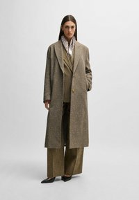 Olive green long coat with a textured, speckled finish; worn over a check-patterned suit. Black pointed-toe shoes and a light scarf complete the look.