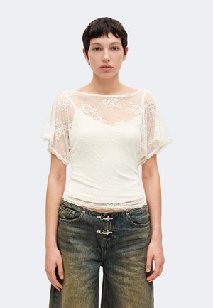 Jeune personne aux cheveux courts et foncés porte un haut blanc en dentelle à manches courtes et un jean délavé et ample avec des fermetures à double boucle uniques.