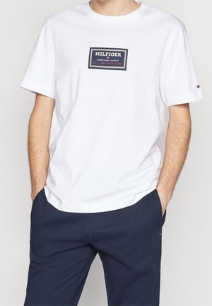 T-shirt en coton blanc avec un patch logo rectangulaire bleu marine et rouge. Manches courtes et encolure ronde. Texture lisse.