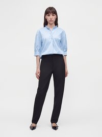 Femme debout portant un chemisier bleu clair à manches trois-quarts, un pantalon noir taille haute et des ballerines noires pointues.