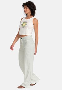 Hvit kort tanktopp med grafisk design kombinert med lysegrønne høyt midje flared jeans og brune sandaler. Glatt stoff og avslappet passform.