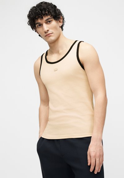 Jeune homme aux cheveux bouclés portant un débardeur beige avec des bordures noires et un pantalon sombre, debout avec une main dans la poche.