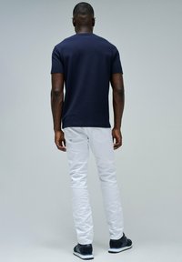 Hombre de pie de espaldas, vistiendo una camisa corta azul marino, pantalones blancos y zapatillas azul marino frente a un fondo liso de color gris claro.