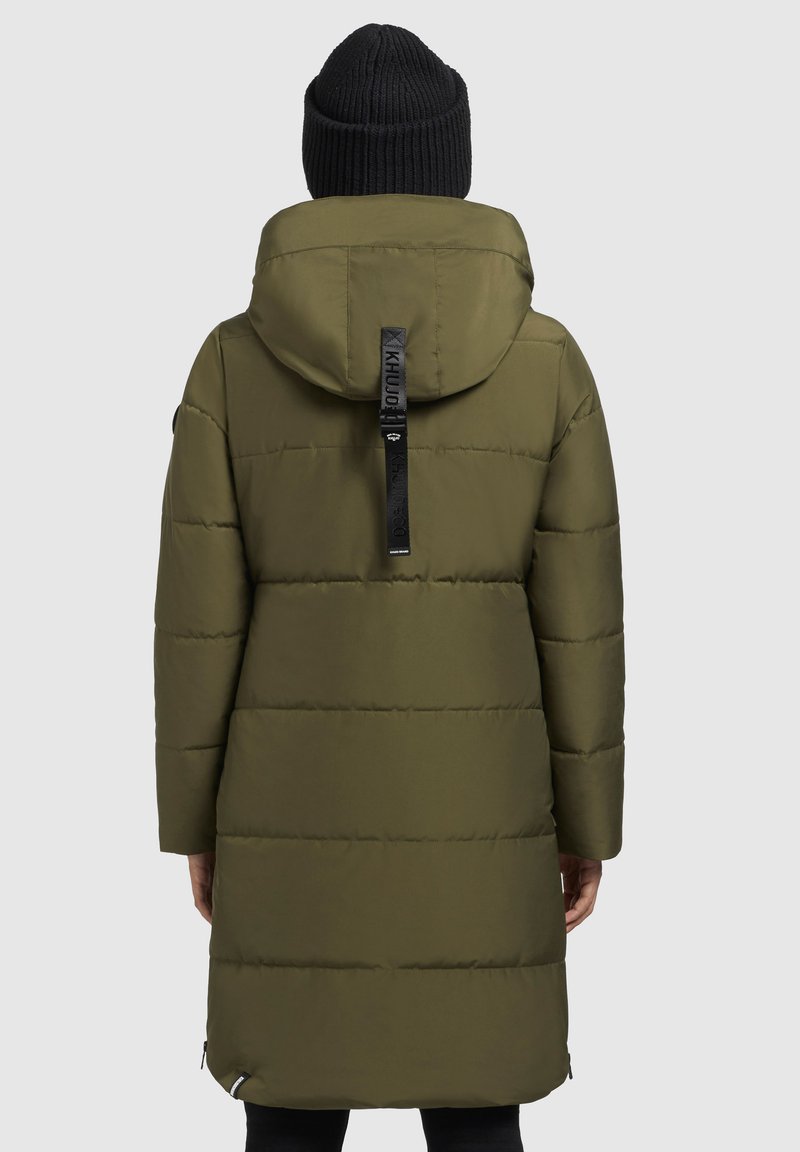 Olivgrüne Steppjacke mit Kapuze, mit einem Logo-Aufnäher auf dem Rücken, vertikalen Nahtlinien und einem knielangen Design. Schwarze Strickmütze.