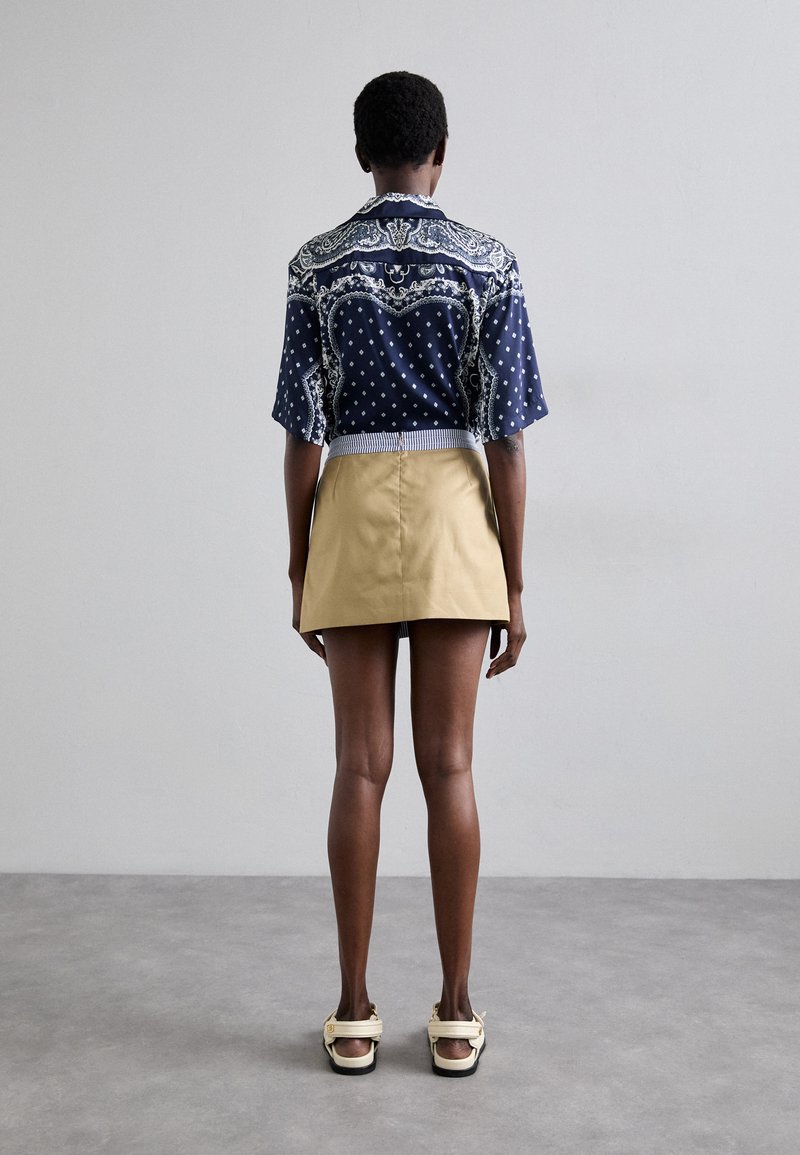 Marineblaues Kurzarmhemd mit weißen Punkten und Paisley-Muster, kombiniert mit einem khakifarbenen Rock. Verfügt über einen gestreiften Bund und lässige Sandalen.