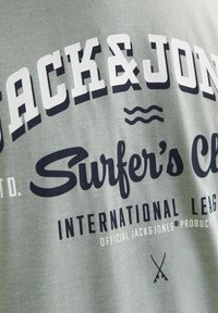 Lys grøn bomulds t-shirt med fedt marineblåt grafisk tekst, herunder "JACK & JONES" og "Surfer's Club", samt subtile tekstur- og designdetaljer.