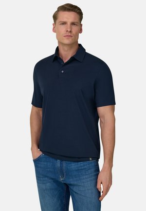 Mężczyzna ubrany w granatową koszulkę polo z krótkim rękawem i niebieskie dżinsy, stoi z jedną ręką w kieszeni na jednolitym tle.