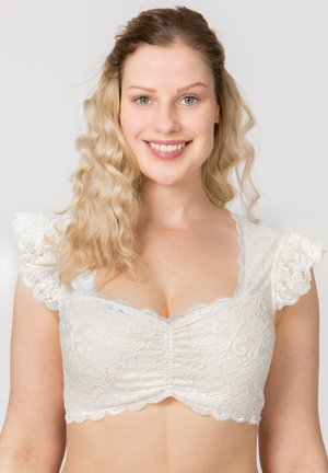 Lächelnde Frau mit welligem blondem Haar, die ein weißes Spitzen-Crop-Top mit gerüschten Kappärmeln trägt, vor einem schlichten hellen Hintergrund.